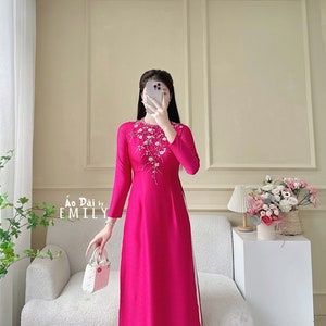 May include: A pink long-sleeved dress with white floral embroidery on the front. The dress has a side slit and is paired with white pants. The text "Ao Dai by EMILY" is visible on the dress.