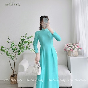 May include: A turquoise long-sleeved dress with a white panel down the side and pearl accents. The dress has a mandarin collar and is made of a silky fabric. The dress is being modelled by a person holding a white handbag.