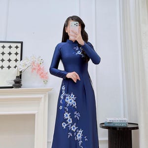 May include: A floor-length navy blue Ao dai dress featuring long sleeves, a high collar, and white floral embroidery with blue accents on the chest and side. The garment is worn over white trousers and includes the text Ao dai Emily at the bottom.