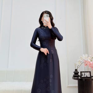 Könnte beinhalten: Ein bodenlanges dunkelblaues Kleid mit langen Ärmeln und einem hohen Kragen. Das Kleidungsstück weist ein dezentes, tonales Blumenmuster auf dem gesamten Stoff sowie eine dekorative Perlenverzierung am Ausschnitt auf. Das Kleid hat ein enges Oberteil.