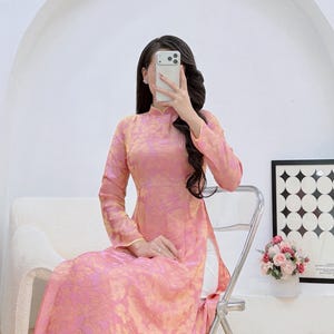 May include: Pink and gold floral patterned Ao dai dress with long sleeves and a mandarin collar. The dress is worn with white pants. The text "Ao dai Emily" is visible on the bottom left of the image.