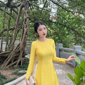 May include: A woman wearing a long-sleeved yellow dress with intricate embroidery around the neckline. The dress has a high collar and a flowing skirt. The text "Ao Dai Emily" is visible on the right side of the image. She is holding a small, striped clutch.