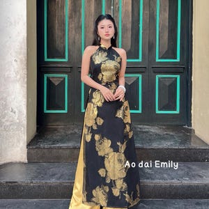 May include: A black halter-neck dress with gold floral patterns and a gold underskirt. The dress is worn in front of a dark wooden door with teal trim. The text "Ao dai Emily" is visible on the dress.