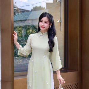 Puede incluir: Un vestido Aodai verde claro con detalles blancos y adornos florales. El vestido tiene cuello mandarín y mangas tres cuartos. El sitio web AodaiEmily.com es visible en el vestido. Se sostiene un pequeño bolso blanco.