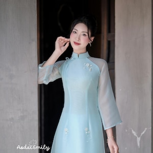 Puede incluir: Un vestido Áo dài vietnamita azul claro con mangas transparentes y adornos florales. El vestido presenta un cuello alto y está adornado con delicados detalles florales blancos. Es una prenda tradicional vietnamita.