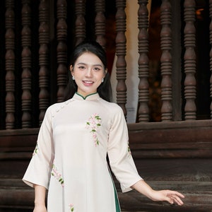Puede incluir: Una mujer lleva un vestido largo blanco con bordado floral rosa y ribete verde. Lleva un bolso de paja con un adorno de flor blanca. El vestido tiene un cuello mandarín y cierres de botón. El texto "Áo Dài by EMILY" es visible en la esquina inferior derecha de la imagen.