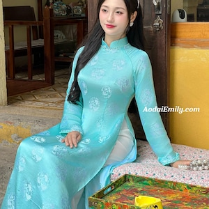 May include: A light blue Vietnamese Áo dài dress with floral patterns. The long-sleeved dress has a mandarin collar and a high side slit. The website AodaiEmily.com is visible on the dress. The model is sitting on a bench with a colourful tray in front.