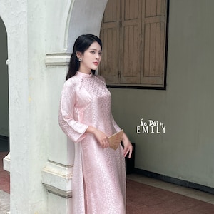 Könnte beinhalten: Ein rosa Seidenkleid mit Stehkragen und langen Ärmeln. Das Kleid hat ein Blumenmuster und einen seitlichen Schlitz. Das Kleid wird von einer Frau getragen, die vor einem weißen Torbogen steht. Der Text "Áo Dài by EMILLY" ist auf dem Kleid sichtbar.