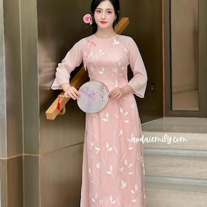 Puede incluir: Un vestido rosa claro con bordados florales blancos y mangas largas y transparentes. El vestido presenta un cuello alto con un detalle de lazo y una falda fluida. Un abanico redondo y decorativo se sostiene frente al vestido. El sitio web es visible.