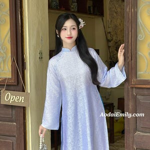 May include: A light blue ao dai dress with a floral pattern. The dress has long sleeves and a mandarin collar. The website address AodaiEmily.com is visible. A small sign that says "Open" hangs on the door.