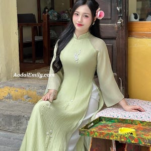 Op de afbeelding: Een lichtgroene Aodai-jurk met lange mouwen en een hoge kraag. De jurk heeft bloemversieringen en een split aan de voorkant, gecombineerd met een witte broek. Het websiteadres AodaiEmily.com is zichtbaar. Een roze bloem siert het haar.