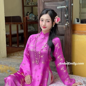 May include: A vibrant pink Aodai dress with gold floral patterns. The long-sleeved dress features a mandarin collar and is accessorized with a pearl necklace. The website address AodaiEmily.com is visible on the right side of the image.