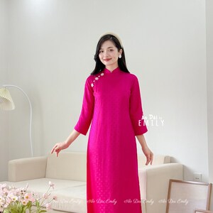 Puede incluir: Un vestido rosa de manga larga con cuello mandarín y bordado floral en el hombro. El vestido se combina con un pantalón rosa y zapatos blancos. El texto "Ao Dai by EMILY" es visible en el vestido.