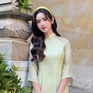 Op de afbeelding: Een vrouw in een lichtgroene traditionele Vietnamese jurk met witte borduursels en een groen hoofdband. Ze houdt een witte parelhandtas vast. De jurk heeft een transparante overlay op de mouwen. De tekst "Ao Dai Emily" is zichtbaar in de rechterbenedenhoek van de afbeelding.