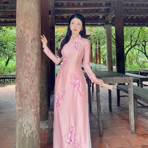 May include: A long, pink dress with floral embroidery. The dress has long sleeves and a mandarin collar. The woman is standing outside in a wooden structure. She is holding a fan.