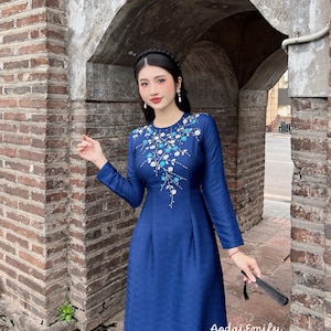 Peut inclure: Une longue robe bleu marine avec broderie florale autour de l'encolure. La robe présente une silhouette fluide et est accessoirisée d'un bandeau noir et de boucles d'oreilles. Le texte "Aodai Emily" est visible en bas à droite.