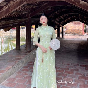 Puede incluir: Una mujer vistiendo un vestido tradicional vietnamita verde claro con bordados florales blancos. Ella sostiene un abanico plateado y está de pie debajo de un puente de madera.