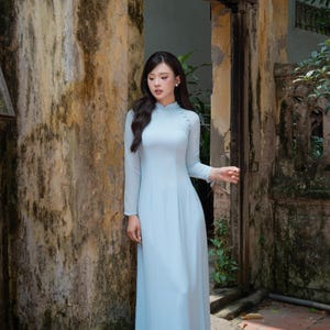 May include: A light blue Ao dai featuring a high collar, long sleeves, and a row of small matching buttons along the side closure. The garment is a floor-length, fitted tunic with a smooth, solid-colored fabric finish and a clean, minimalist design.
