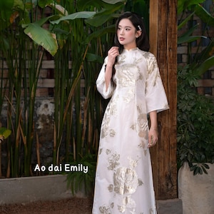 Può includere: Abito Ao dai bianco con ricami floreali dorati e maniche ampie. L'abito è abbinato a pantaloni dorati. Il testo "Ao dai Emily" è visibile nell'immagine. È un capo tradizionale vietnamita.