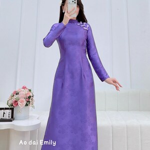 May include: A long, purple dress with a high neckline and long sleeves. The dress features a floral pattern and is adorned with small floral embellishments on the shoulder. The text "Ao dai Emily" is visible at the bottom of the image.