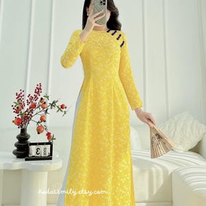 May include: A yellow long-sleeved dress with a floral pattern and a mandarin collar. The dress has a side slit and is worn with a white underskirt. The dress is shown on a model holding a fan.