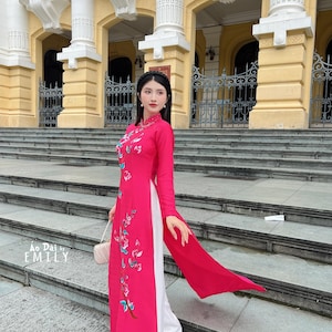 Könnte beinhalten: Eine Frau trägt ein traditionelles vietnamesisches Kleid in Rosa und Weiß, das Ao Dai genannt wird. Das Kleid ist mit Blumen bestickt und hat einen langen, fließenden Rock. Die Frau steht auf einer Treppe vor einem Gebäude mit verzierter Architektur. Der Text "Ao Dai by EMILY" ist in der linken unteren Ecke des Bildes sichtbar.