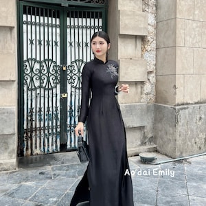 May include: A long, black dress with long sleeves and a high collar. The dress features a floral embroidery detail on the chest and a high slit on the side. The text "Ao dai Emily" is visible on the bottom right of the image. The woman is holding a black purse.