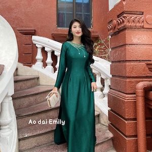 Op de afbeelding: Een lange, smaragdgroene jurk met lange mouwen en een hoge halslijn, versierd met delicate versieringen. De jurk heeft een vloeiend silhouet en wordt gecombineerd met een kleine, crèmekleurige clutch. De tekst "Ao dai Emily" is zichtbaar op de trappen.
