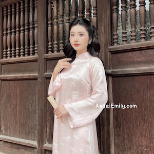 Könnte beinhalten: Hellrosa Ao Dai Kleid mit floralem Muster. Das langärmlige Kleid hat einen Mandarin-Kragen und einen Schlitz an der Seite. Die Website AodaiEmily.com ist auf das Kleid gedruckt.