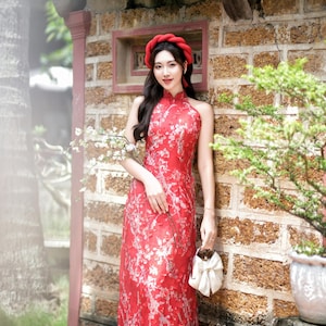 May include: A woman wearing a red dress with white floral embroidery stands in front of a brick wall. She is holding a white clutch purse and has a red headband on. The text "Aodai Emily" is visible in the bottom right corner of the image.