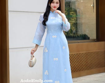 May include: A light blue ao dai dress with white floral embroidery. The dress has a mandarin collar and three-quarter sleeves. The dress is paired with white pants. The website address AodaiEmily.com is visible.