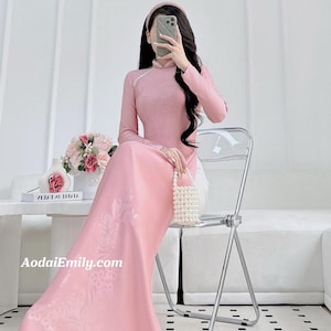 May include: A pink, long-sleeved dress with floral patterns, a mandarin collar, and a matching headband. The dress is floor-length and has a subtle sheen. A small, beaded purse is held. The website address AodaiEmily.com is visible.