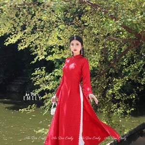 Könnte beinhalten: Ein rotes Ao Dai Kleid mit langen Ärmeln und einem hohen Kragen. Das Kleid hat einen Schlitz vorne, der weißen Stoff darunter zeigt. Die Ärmel und Kanten sind mit kleinen, dekorativen Details verziert. Der Text "Ao Dai Emily" ist sichtbar.