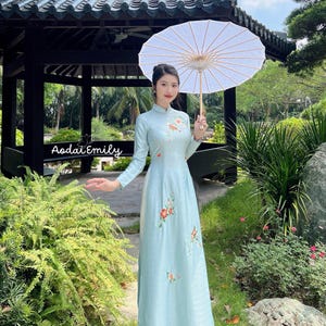 May include: A woman wearing a light blue traditional Vietnamese dress with floral embroidery, holding a white paper parasol in a garden setting.