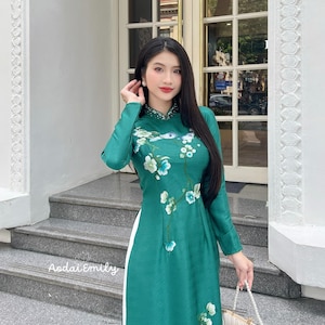 May include: A teal-green Áo dài dress with long sleeves and floral embroidery. The dress features a high neckline adorned with small beads and a white panel down the side. A small, cream-colored purse with a pearl handle is held in the hand.