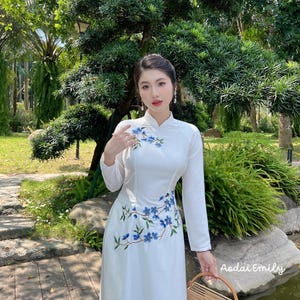 Könnte beinhalten: Ein weißes Kleid mit langen Ärmeln und blauer Blumenstickerei. Das Kleid hat einen Mandarin-Kragen und eine taillierte Passform. Das Model steht in einer Gartenlandschaft mit einem Teich und einem Holzsteg.