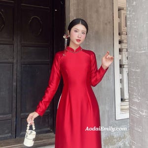 Könnte beinhalten: Ein rotes Kleid mit langen Ärmeln und hohem Ausschnitt. Das Kleid besteht aus einem glatten, glänzenden Stoff und hat ein figurbetontes Oberteil, das in einen weiten Rock übergeht. Eine kleine, weiße, perlenbesetzte Handtasche wird in der Hand gehalten. Die Website AodaiEmily.com ist sichtbar.