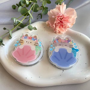 May include: Two clear acrylic phone grips featuring a cartoon cat design. One cat is in a pink seashell, the other in a blue seashell. Both cats wear flower crowns and have mermaid tails. The grips are on a white, oval-shaped dish.