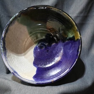 Puede incluir: Un cuenco de cerámica con un esmalte blanco, marrón, verde y morado. El cuenco tiene una forma redondeada y un borde ligeramente acampanado.