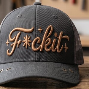 May include: A charcoal grey trucker hat featuring a mesh back and a structured front panel. The front displays the embroidered text F*ckit in a metallic copper script font, accented by small four-pointed star symbols on either side of the lettering.