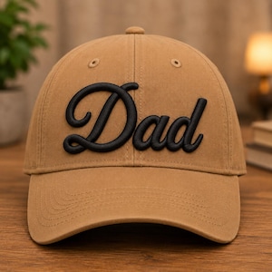 May include: A tan baseball cap featuring the word Dad embroidered in black script lettering across the front panels. The cap has a structured crown, a curved brim, and stitched eyelets on the top panels for ventilation and a classic design.