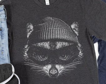 May include: Two t-shirts, one dark gray and one navy blue, each featuring a white graphic of a raccoon wearing a beanie. The raccoon's face is detailed with whiskers and a focused expression. The shirts are casual wear.