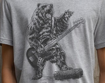 Puede incluir: Una camiseta gris con un gráfico de un oso tocando un bajo. El oso está de pie sobre un tronco y tiene una expresión decidida en su rostro.