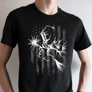 May include: Black t-shirt with a white graphic of a welder wearing a welding mask and holding a welding torch in front of a distressed American flag.