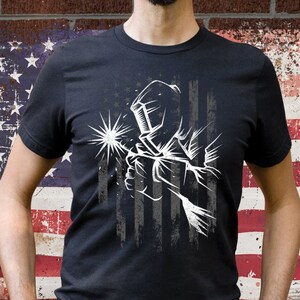 May include: A black t-shirt with a white graphic of a welder wearing a welding mask in front of a distressed American flag design.