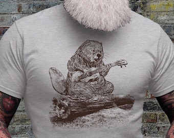Puede incluir: Camiseta gris claro con una ilustración marrón de un castor tocando el ukelele. El castor está representado con pelaje y el ukelele tiene cuerdas. El castor está sentado en un tronco. Prenda casual.