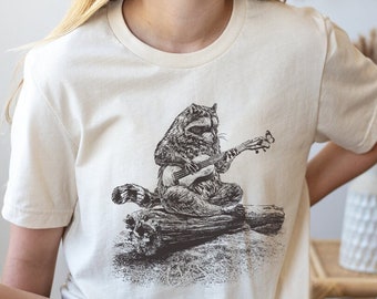 Könnte beinhalten: Cremefarbenes T-Shirt mit einer Schwarz-Weiß-Illustration eines Waschbären, der Ukulele spielt und auf einem Baumstamm sitzt. Der Waschbär ist der zentrale Fokus, mit detaillierten Schattierungen. Das Shirt ist ein lässiger Stil mit kurzen Ärmeln.