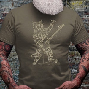 Może przedstawiać: Koszulka w kolorze khaki z grafiką przedstawiającą kota grającego na gitarze basowej. Kot jest w rockowej pozie z otwartymi ustami i uniesionym ogonem. Grafika ma vintage'owy, zużyty wygląd.