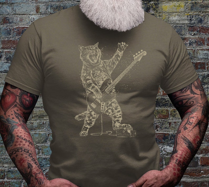 Może przedstawiać: Koszulka w kolorze khaki z grafiką przedstawiającą kota grającego na gitarze basowej. Kot jest w rockowej pozie z otwartymi ustami i uniesionym ogonem. Grafika ma vintage'owy, zużyty wygląd.