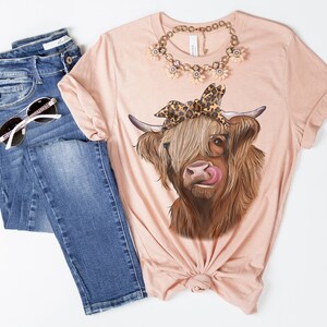 Puede incluir: Una camiseta rosa con una vaca Highland marr&oacute;n y blanca que lleva un pa&ntilde;uelo de leopardo y gafas. La vaca est&aacute; sacando la lengua.